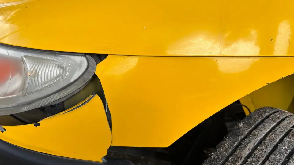 Pare choc avant de voiture rayé et enfoncé, vu de trois quarts avant, dans un atelier de carrosserie.